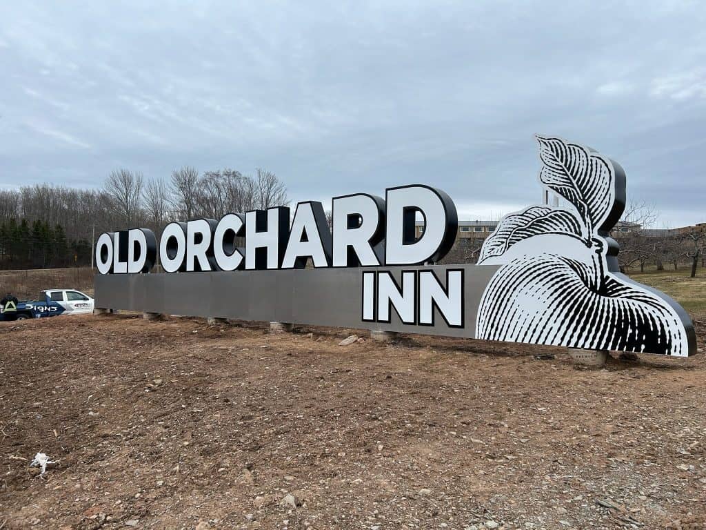Large outdoor sign for Old Orchard Inn featuring bold white 3D letters and a stylized black-and-white apple illustration, installed on a cleared plot of land with a branded Auto Trim & Signs truck visible in the background.