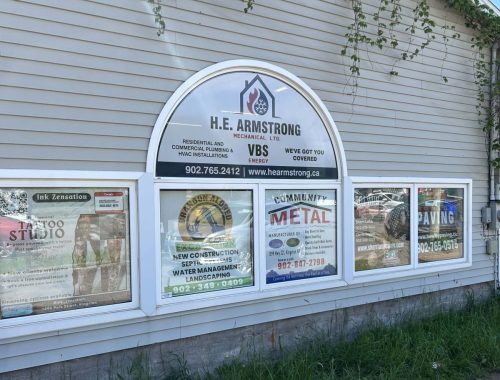Storefront windows displaying multiple business advertisements and logos, including H.E. Armstrong Mechanical, on a commercial building in Kingston, Nova Scotia.