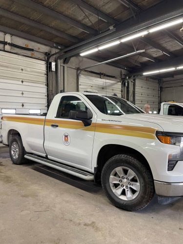 Front side angle of a white municipal Chevrolet Silverado truck with yellow and orange side striping and door logo decals installed inside a garage bay.