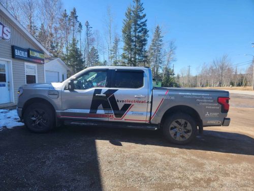 Silver pickup truck wrapped with KVS Electrical Inc. branding, featuring large black and red lettering and service details, parked outside Auto Trim  Signs with signage for backlit and illuminated signs visible in the background.