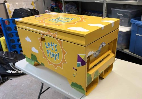 Bright yellow outdoor storage bin wrapped with playful “Let’s Play!” graphics, clouds, kites, and colourful illustrations for a community recreation space.