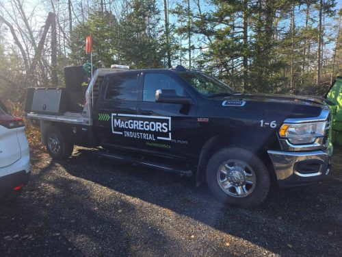 Side view of a black RAM 3500 with MacGregors Industrial decals in white and green, parked in a wooded lot.