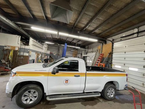 White Chevrolet Silverado with yellow and gold municipal striping and crest installed on the driver-side door inside a garage.