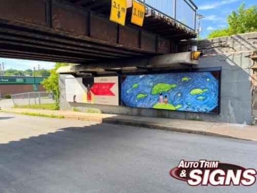 A mural under a bridge features two distinct sections. On the left, there is a bold and graphic banner with the text MMIW and No More Stolen Sisters, depicting a person and a red dress symbolizing missing and murdered Indigenous women. On the right, a colorful painting showcases a serene scene of two people standing on a floating island surrounded by turtles, stars, and swirling blue waters.