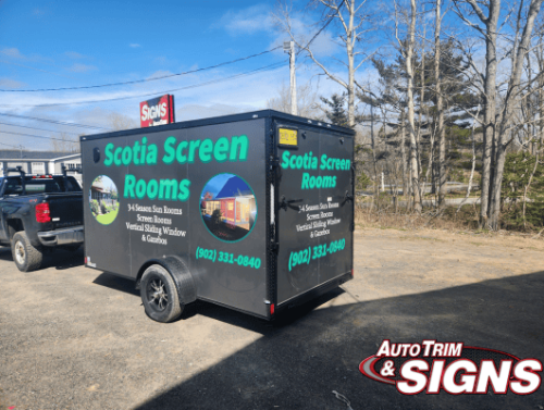 Rear angle view of a black utility trailer parked outdoors, featuring advertising wraps for 'Scotia Screen Rooms.' The graphics showcase images of sunrooms and include contact information. The trailer is parked on a gravel surface with trees in the background under a clear sky