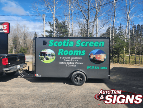 Side view of a black utility trailer parked outdoors with vibrant advertising for 'Scotia Screen Rooms.' The wrap displays a large green logo, images of sunroom projects, and contact details. The setting is a grassy area with a wooded backdrop on a sunny day.