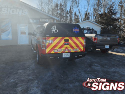 Image of a fire department's pickup truck with a vibrant tailgate wrap featuring yellow and red chevron patterns, a 'Keep Back 150m' safety message, and the fire service logo, parked in front of Auto Trim  Signs shop