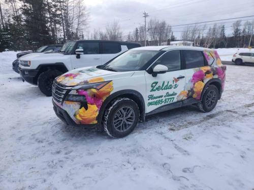 A white Nissan Rogue wrapped with a vibrant floral design for Zelda’s Flower Studio, featuring colorful flowers on the hood and sides, parked outside in the snow.