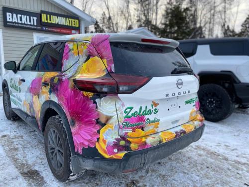 The back view of Zelda’s Flower Studio wrapped Nissan Rogue, showcasing bright floral graphics and business branding, parked outside a signage shop in the snow.