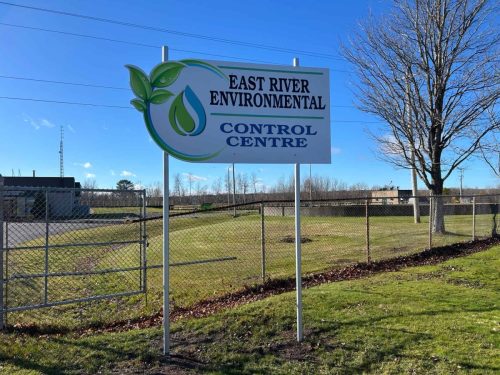 Freestanding outdoor sign for East River Environmental Control Centre installed on a grassy area beside a chain-link fence under a bright blue sky.