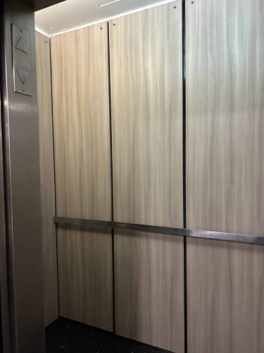 Elevator interior wrapped in woodgrain vinyl with clean panel finish and metal handrail
