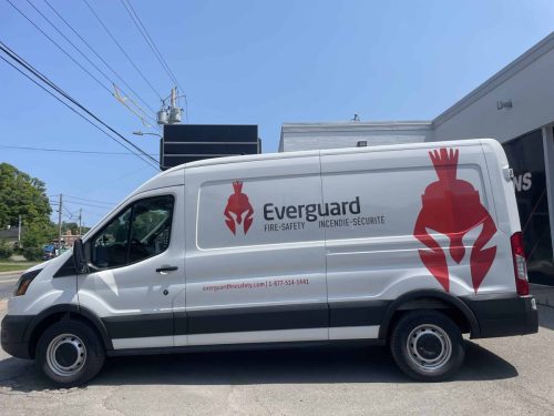 Full side view of white Everguard Fire Safety van with large red helmet graphic, company logo, bilingual text, website, and phone number.