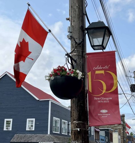 Red pole banner with bold gold “150” and “Celebrate! New Glasgow 1875–2025” text, mounted beside a Canadian flag and flower basket.