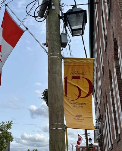 Yellow New Glasgow 150 banner with “Celebrate! 150” and town branding, mounted on a utility pole with Canadian flags in the background.