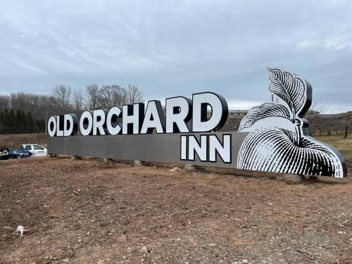 Large outdoor sign for Old Orchard Inn featuring bold white 3D letters and a stylized black-and-white apple illustration, installed on a cleared plot of land with a branded Auto Trim  Signs truck visible in the background.