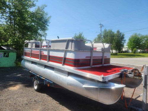 Front and side view of a Princecraft pontoon boat with red flooring and aluminum pontoons, parked outdoors on a trailer under a clear blue sky.