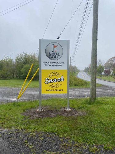 Street-side view of a tall sign displaying Simply Golf and Big Cove Snack Market, installed on a grassy area beside a rural road.