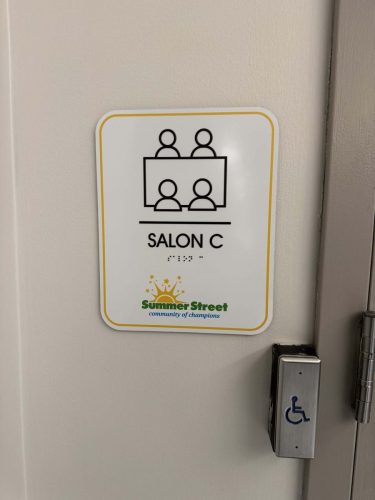 Accessible room identification sign for Salon C at Summer Street featuring braille, meeting room iconography, and rounded-corner design mounted beside a doorway.