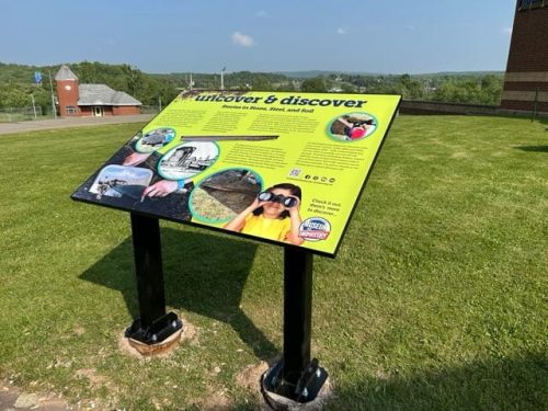Outdoor interpretive sign titled “Uncover  Discover: Stories in Stone, Steel, and Soil” on a grassy lawn with historical photos and a child wearing binoculars.