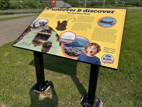 River-themed display titled “Uncover  Discover: The East River Flows” showing images of boats, a scenic river, and a surprised child, set on a grassy landscape.