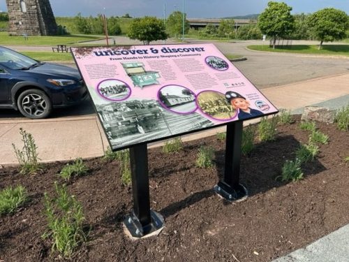 Pink-accented interpretive sign titled “Uncover  Discover: From Hands to History: Shaping a Community” near a parking area, featuring historical photos of people, buildings, and a woman in uniform.