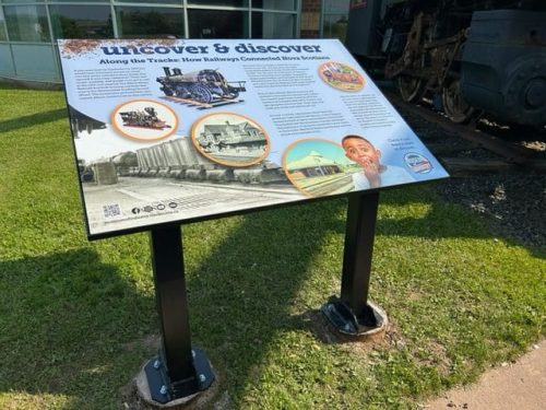 Railway-themed educational sign titled “Uncover  Discover: Along the Tracks: How Railways Connected Nova Scotia,” placed near a historical train.