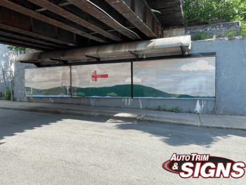 A mural under a bridge shows a panoramic landscape. The painting includes a rolling green hill in the foreground, a serene river at the base, and a sky with soft pastel colors and white clouds. A red cross symbol is prominent in the middle, adding a focal point to the tranquil scene.